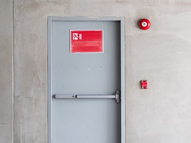 Graue Brandschutztür mit Fluchtwegsymbol und roter Alarmglocke.